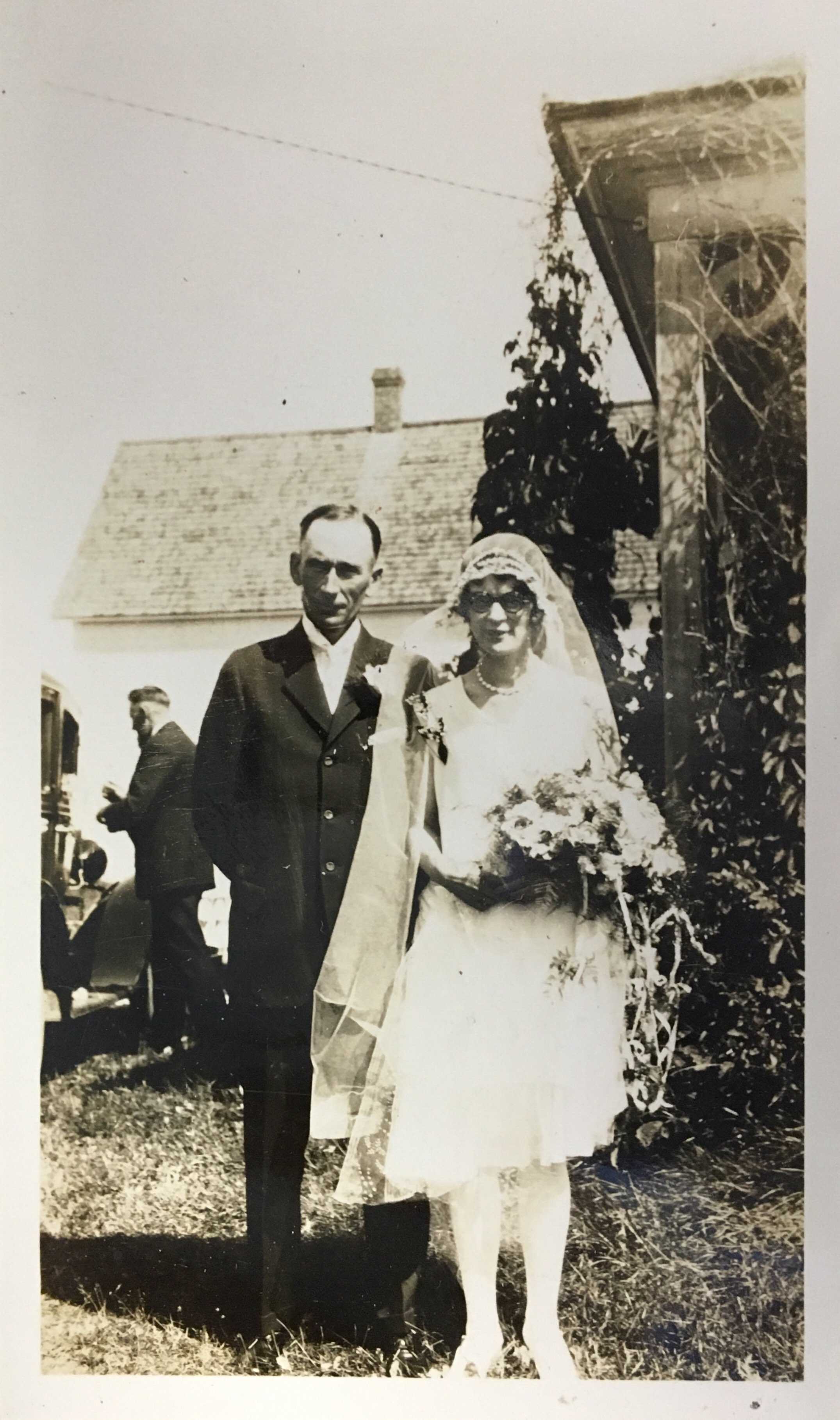 Black and white photograph. Archie and Grace pose for the camera after their wedding. Archie wears a suit, and Grace is in a short gown and veil, holding a large bouquet of flowers.